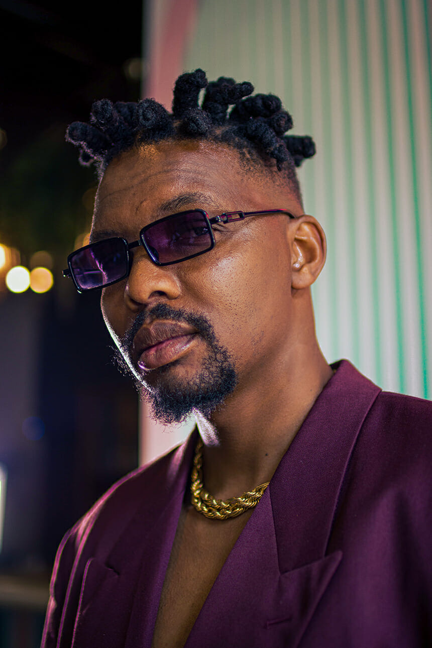 A man with short, twisted hair and a goatee wears dark sunglasses, a chunky gold chain, and a maroon suit jacket with no shirt. He poses confidently in a stylish, well-lit setting with blurred lights and vertical lines in the background.