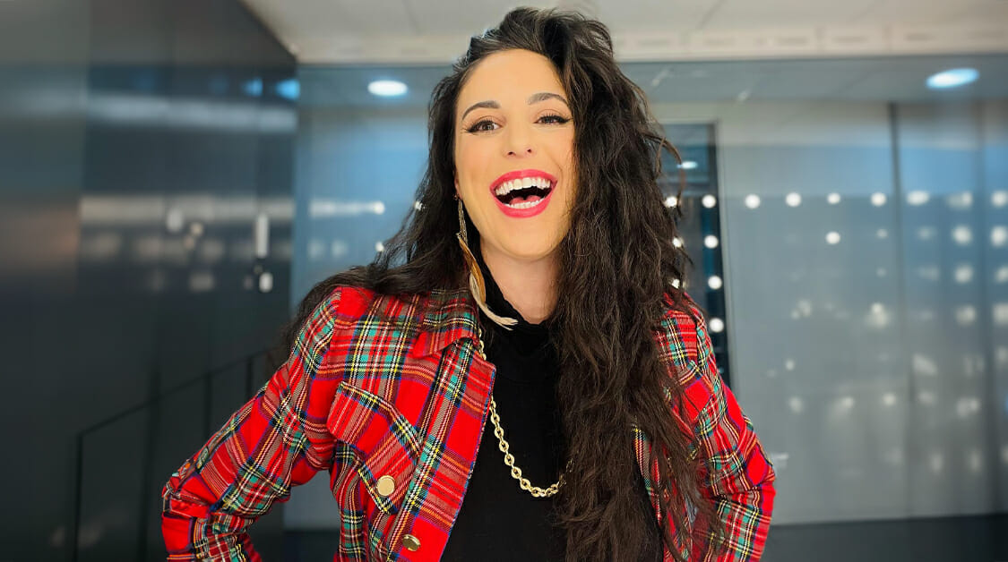 A woman with long wavy dark hair smiles widely. She wears a red plaid jacket over a black top, accessorized with a gold chain necklace. The background is modern and blurred, with reflective panels and circular lights.