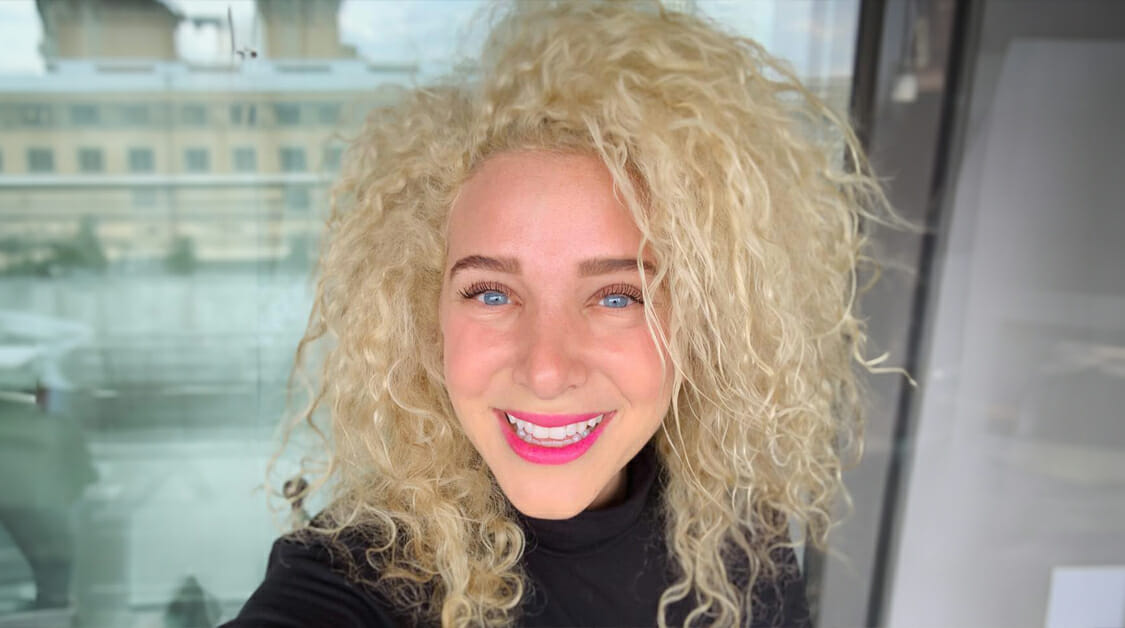 A person with light skin, bright blue eyes, and voluminous curly blonde hair smiles at the camera. They wear a black top and pink lipstick, standing in front of a glass window with buildings faintly visible outside.