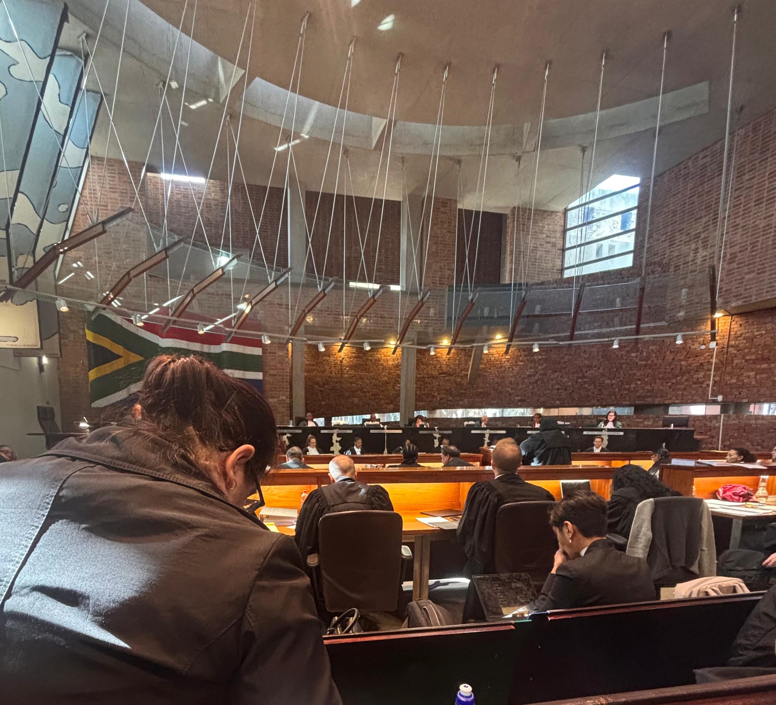 A courtroom with brick walls and a curved ceiling, filled with people seated at desks. Sunlight streams through high windows. A large South African flag hangs on the left. The atmosphere is formal, with attendees focused on proceedings.
